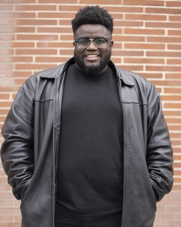 Man with glasses and a beard wearing a black sweater and a gray leather jacket, standing in front of a brick wall smile