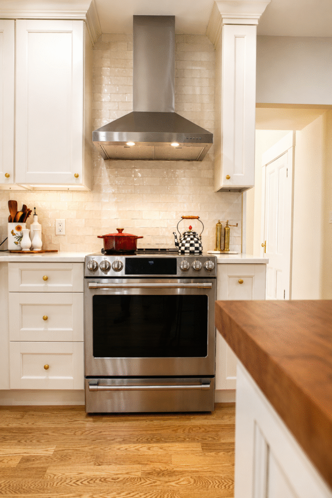 Stainless steel gas range with a range hood in a bright white kitchen, a red pot on the stove and a checkered kettle on the counter.